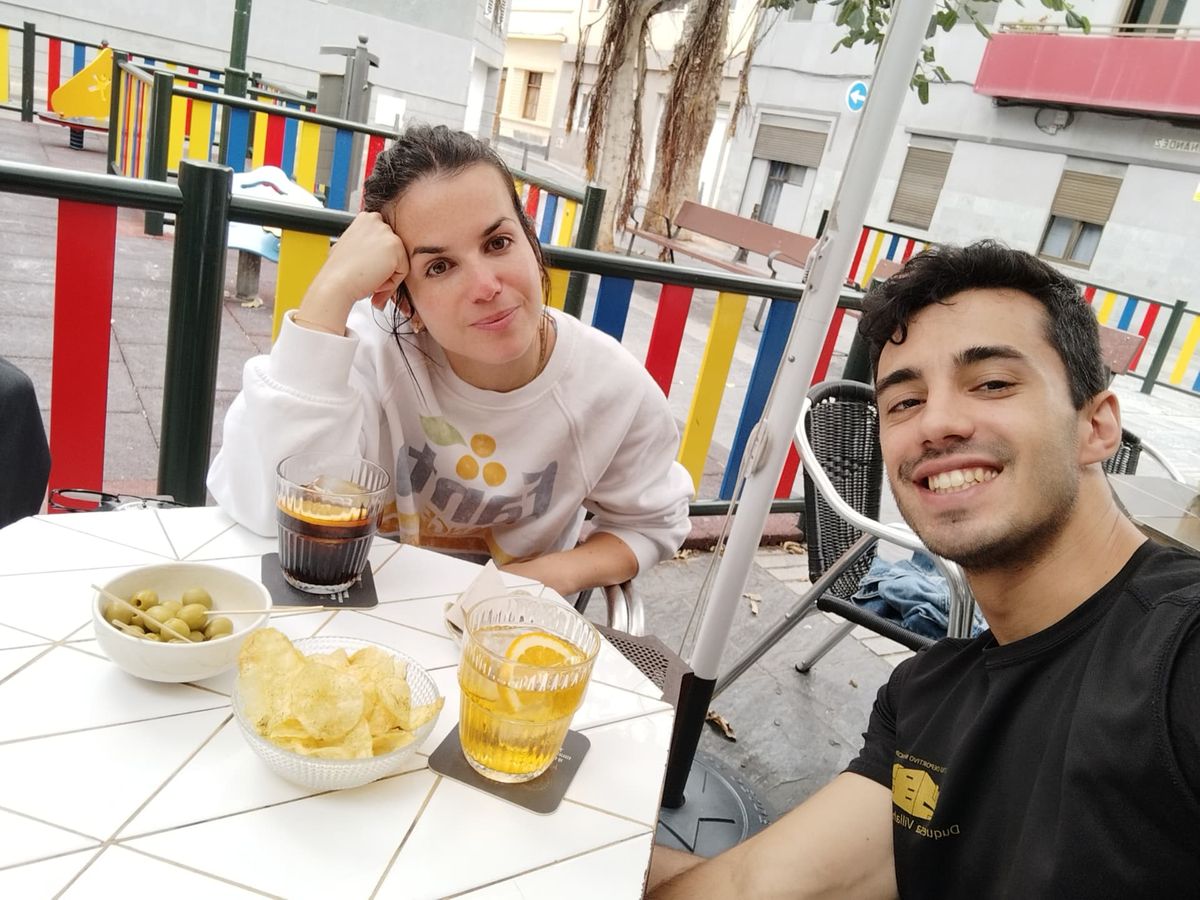 David y Lucía sentados en una terraza, con bebidas y unos aperitivos en la mesa.