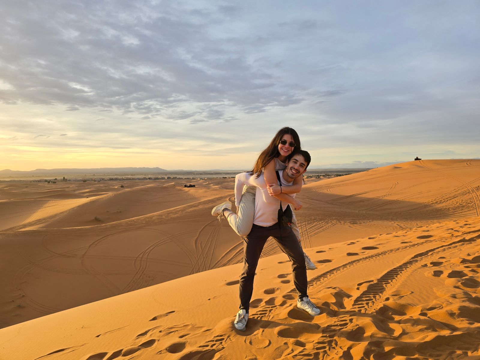David y Lucía sonriendo sobre una duna del desierto al atardecer; Lucía va subida a la espalda de David.